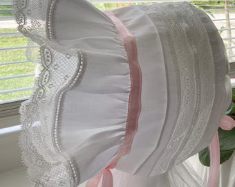 May include: A white bonnet featuring delicate lace trim along the ruffled brim and crown. The headwear is accented with a soft pink ribbon woven through the fabric, which also forms ties at the bottom to secure the bonnet under the chin.