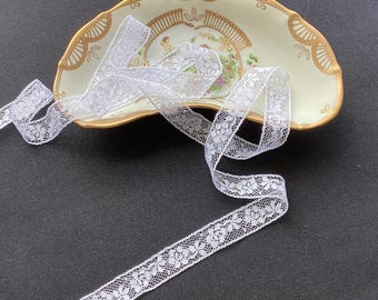 May include: A length of white floral lace ribbon draped over a crescent-shaped ceramic dish. The dish features a cream-colored base with gold-painted scalloped edges and a central illustration of cherubs surrounded by green and pink floral accents.