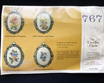 May include: A Creative Circle cross-stitch kit package labeled 767, featuring four oval designs: Summer Bouquet, Stocks and Tulips, Blue Bird, and Morning Glories. The package includes 100% cotton 22-count Hardanger cloth, embroidery floss, a die-cut mounting board, a cast metal frame with an easel back, a needle, a graph, and instructions. The finished size is 2.5 inches by 3.5 inches.
