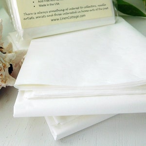 May include: A stack of white, folded fabric sheets, possibly linen, are the primary focus. A seashell and a package with text are in the background. The text on the package includes "Acid Free" and "Made in the USA". The website address is www.LinenCottage.com.
