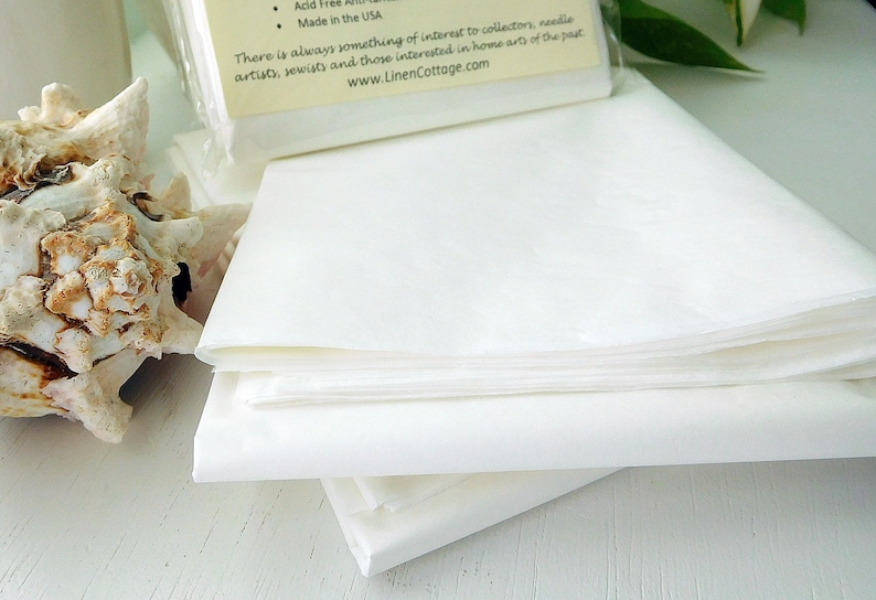 May include: A stack of white, folded fabric sheets, possibly linen, are the primary focus. A seashell and a package with text are in the background. The text on the package includes "Acid Free" and "Made in the USA". The website address is www.LinenCottage.com.