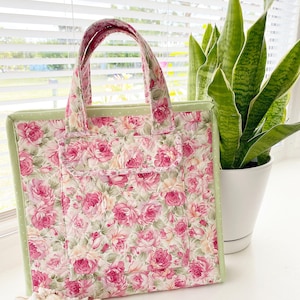 May include: A floral tote bag with a pink rose pattern. The bag has a light green border and handles, and a front pocket. The bag is sitting next to a potted plant and seashells.