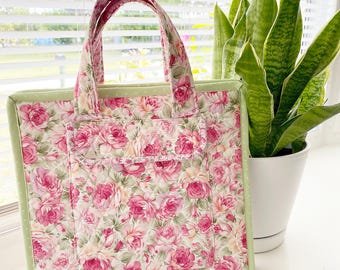 May include: A floral tote bag with a pink rose pattern. The bag has a light green border and handles, and a front pocket. The bag is sitting next to a potted plant and seashells.