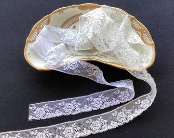 May include: A length of white floral lace trim with a scalloped edge, coiled and draped over a semi-circular ceramic dish featuring a cream-colored base, gold-painted scalloped edges, and a subtle floral pattern in the center of the dish.