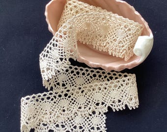 May include: A length of cream-colored crocheted lace trim with a repeating circular pattern and a scalloped edge, coiled inside a pale pink ceramic dish with a shell-like shape and a smooth, glossy finish.