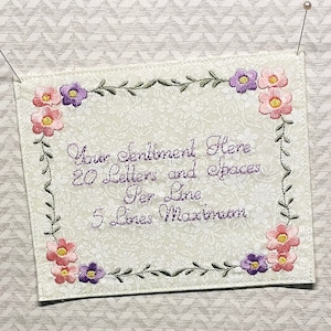 May include: Embroidered cream-colored fabric with floral and vine border. The corners feature pink and purple flowers. Text in the center reads "Your Sentiment Here, 20 Letters and Spaces Per Line, 5 Lines Maximum."