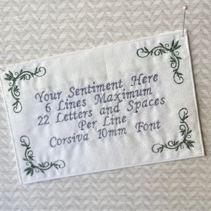 May include: White rectangular fabric with embroidered text: "Your Sentiment Here, 6 Lines Maximum, 22 Letters and Spaces Per Line, Corsiva 10mm Font." Green floral corner designs. Two pins secure the fabric.