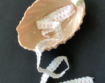 May include: A pale peach-colored ceramic dish shaped like a leaf with a rolled edge, containing a length of white lace trim woven with a thin, light pink ribbon. The lace features a scalloped edge and a repeating pattern of small pink squares.