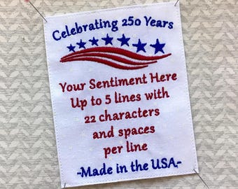 May include: White embroidered fabric with text in blue and red. The text reads "Celebrating 250 Years" above a design of stars and stripes. Below, it says "Your Sentiment Here" and details for customization. The bottom reads "Made in the USA".