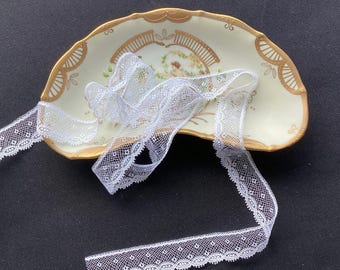 May include: A crescent-shaped ceramic dish with a cream-colored center, gold-painted scalloped edges, and a central illustration of a figure in a garden. A length of white, delicate, patterned lace ribbon is draped across the dish.