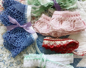 May include: A collection of decorative lace and fabric trims including a blue crocheted piece tied with a purple ribbon, a light green lace trim, a pink crocheted ruffle, a red and white patterned lace trim, and a white fabric strip with green stitching.