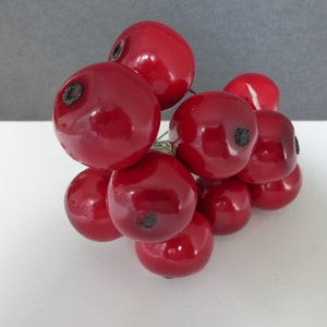 May include: A cluster of glossy, deep red artificial berries. Each berry has a small, dark indentation at the top. The berries are arranged together, suggesting they are part of a decorative floral arrangement or craft supply.
