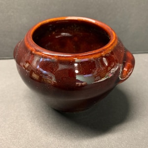 May include: A small, brown ceramic pot with two handles. The pot is glazed and has a shiny finish.