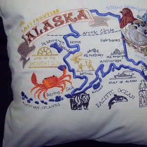 May include: Embroidered pillow featuring a map of Alaska with various landmarks and animals, including a moose, a bear, a crab, a bird, and a whale. The text "The Last Frontier Alaska" is embroidered at the top of the pillow.