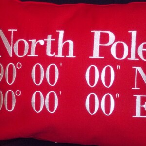May include: A red rectangular pillow with white embroidered text. The text reads "North Pole" above the coordinates 90&deg; 00' 00" N and 00&deg; 00' 00" E. The pillow is a solid red color.