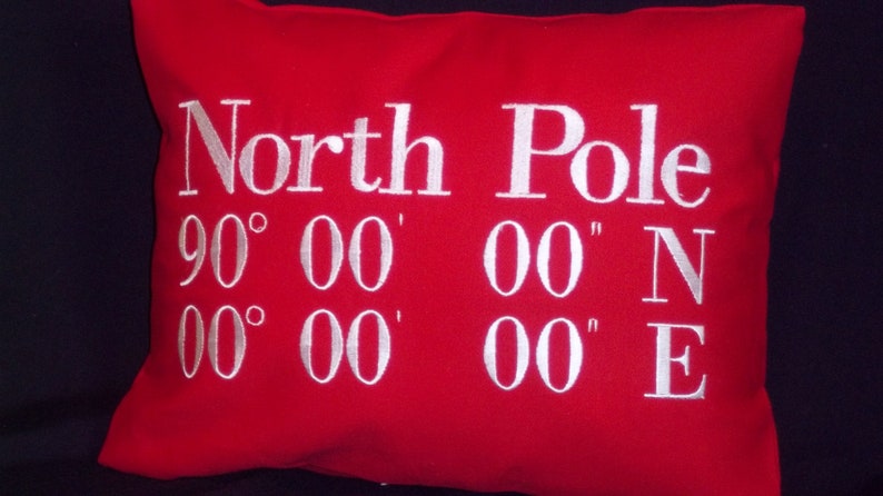 May include: A red rectangular pillow with white embroidered text. The text reads "North Pole" above the coordinates 90&deg; 00' 00" N and 00&deg; 00' 00" E. The pillow is a solid red color.