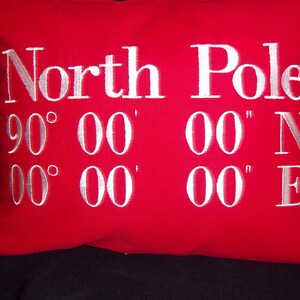May include: A red decorative pillow with white embroidered text that reads "North Pole" and the coordinates 90&deg; 00' 00" N and 00&deg; 00' 00" E. The pillow is a square shape.