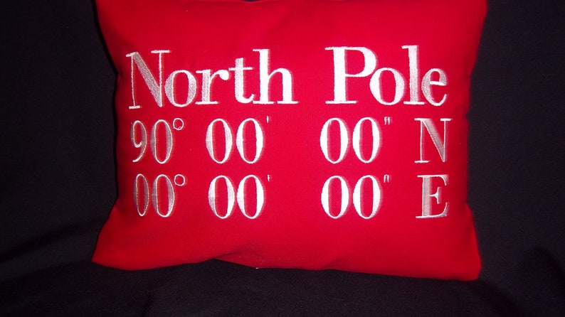 May include: A red decorative pillow with white embroidered text that reads "North Pole" and the coordinates 90&deg; 00' 00" N and 00&deg; 00' 00" E. The pillow is a square shape.