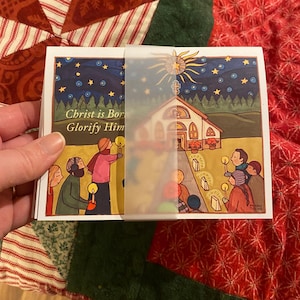 May include: A Christmas card with a colorful illustration of a church, a star, and people holding candles. The text on the card reads "Christ is Born, Glorify Him".