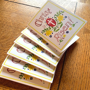 May include: A stack of white greeting cards with a yellow border. The top card has a red and white cross with the text "Christ is Risen!" in red and black lettering with yellow and blue flowers.