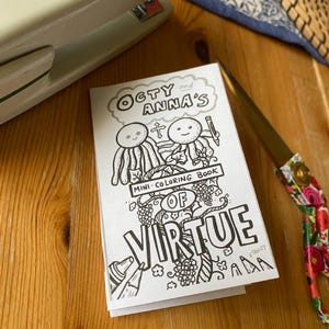 May include: A mini coloring book titled "Octy and Anna's of Virtue" with black and white illustrations. The book is open on a wooden surface, next to a stapler and floral scissors.