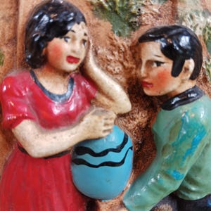 May include: A ceramic relief sculpture depicting two figures, a person in a red dress and another in a green shirt, holding a blue ball with black stripes. The figures have dark hair and are set against a textured, brown background.