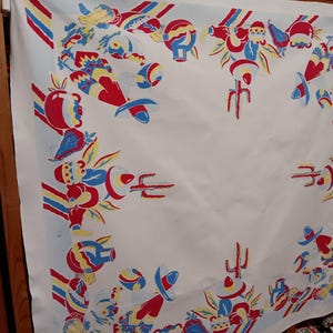 May include: A square white tablecloth featuring a repeating border print of red, blue, and yellow illustrations. The pattern includes depictions of sombreros, cacti, pottery, and chili peppers arranged in a decorative frame around the center.