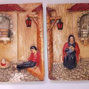 May include: Two-piece wall art depicting a Mexican village scene. One piece shows a man in a red shirt and black pants resting, with a sombrero nearby. The other shows a woman in a red shawl. Both pieces have red tile roofs and arched windows.