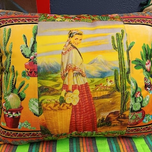 May include: A yellow throw pillow with a colorful embroidered design featuring a woman in a traditional dress, cacti, and a basket of flowers. The pillow has green pom-poms around the edges.
