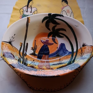 May include: Oval ceramic serving bowl with a hand-painted desert scene featuring a figure in a sombrero, cacti, and palm trees. The bowl has a wide rim and a stitched leather trim. The background includes a yellow fabric with two painted figures.