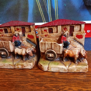 May include: A pair of bookends made of painted ceramic. Each bookend features a small, red-roofed building with a person in a red shirt and a straw hat standing in front of a small cart pulled by two oxen. The bookends are on a wooden surface.