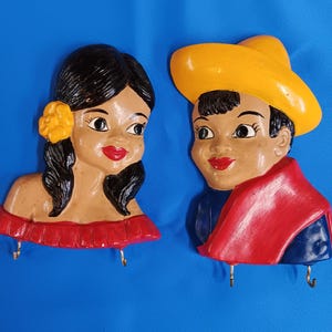 May include: A pair of ceramic wall hooks featuring a woman and a man in traditional Mexican attire. The woman is wearing a red dress with a yellow flower in her hair. The man is wearing a yellow sombrero and a red scarf. Both figures have painted details on their faces.
