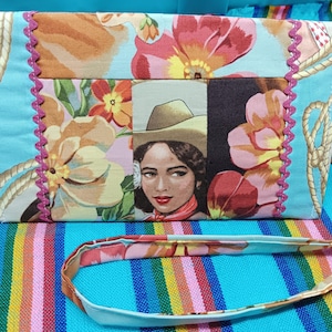 May include: A rectangular fabric crossbody bag featuring a light blue background with a floral pattern, rope motifs, and a central panel depicting a person wearing a cowboy hat. The bag is accented with pink zigzag trim and a matching strap.