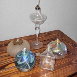 May include: A collection of glass oil lamps in various shapes and sizes. The lamps include a tall, stemmed lamp, a frosted round lamp, a swirled blue and green lamp, a square lamp, and an iridescent round lamp. Each lamp has a wick.