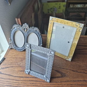 May include: Three decorative picture frames: one dark gray oval double frame with ornate scrollwork, one rectangular gold frame with a textured finish, and one small square silver-toned frame featuring a detailed pattern and embedded clear crystals.