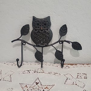 May include: A black metal wall hook featuring an owl perched on a branch with leaves. The hook has three hooks and is designed for hanging items. The owl has detailed features and is a decorative piece for home decor.