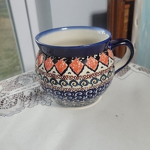 May include: A ceramic mug with a blue rim and handle, featuring a detailed pattern of orange, black, and blue geometric shapes and swirls. The mug has a rounded body and a white interior, resting on a white surface.