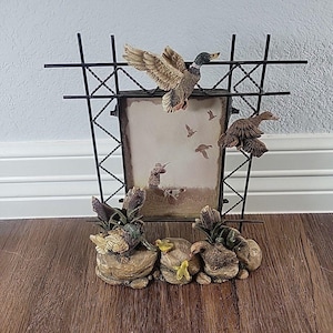 May include: Decorative picture frame with a hunting theme. The frame has a black metal grid design and features sculpted ducks in flight and resting on rocks. The picture inside depicts a hunter and dogs in a field.