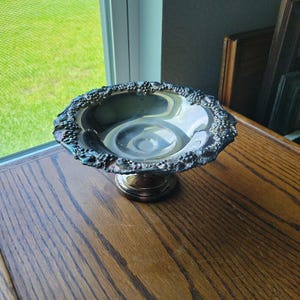 May include: A silver-toned pedestal bowl featuring a polished, reflective interior basin and a decorative rim embossed with a detailed pattern of grape clusters and leaves. The bowl is supported by a circular, raised base, creating a raised display.