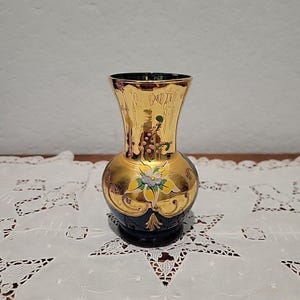 May include: A decorative vase with a gold exterior and a dark blue base. The vase features floral and grape designs in gold, blue, and green. The vase has a rounded body and a flared top, suitable for displaying flowers.