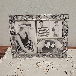 May include: Silver-toned metal multi-photo frame with rose and ribbon detailing. The frame holds four black and white photos of people. The frame is sitting on a white lace tablecloth.