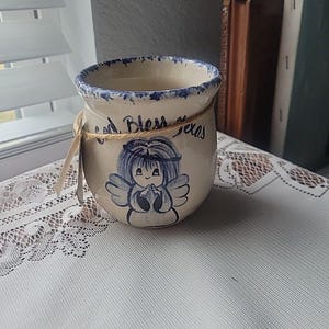 May include: A cream-colored ceramic jar with a blue rim and a hand-painted angel design. The jar has the words "God Bless Texas" and is tied with twine. A small tag hangs from the twine.