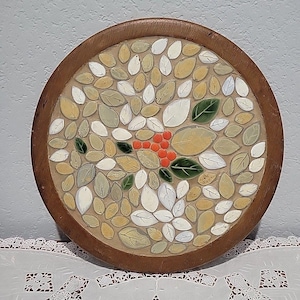 May include: Round wooden trivet with a mosaic design. The mosaic features a pattern of leaves in shades of white, green, and tan, with a cluster of orange berries in the center. The trivet has a dark brown wooden frame.