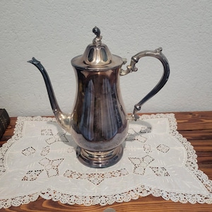 May include: A silver-colored antique coffee pot with a curved spout and handle. The pot has a decorative lid with a small finial. It sits on a white lace doily. The pot is likely made of silver or a silver-plated metal.