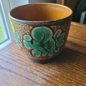 May include: A ceramic planter with a brown exterior and a contrasting orange interior. The planter features embossed green leaf and swirl designs. The rim is dark brown, and the planter sits on a wooden surface.