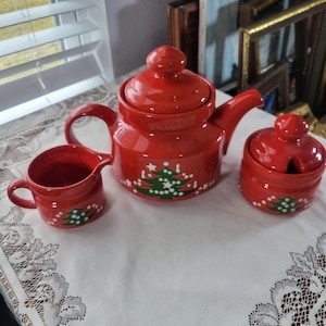 May include: A red ceramic tea set comprising a teapot, creamer jug, and sugar bowl. Each item features a green Christmas tree motif. The set is displayed on a white tablecloth.