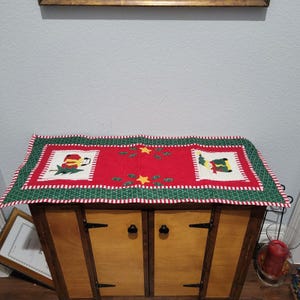 May include: A rectangular Christmas table runner with a red center and green and white striped border. The red center features embroidered images of Santa Claus and an elf, along with stars and sprigs of holly.