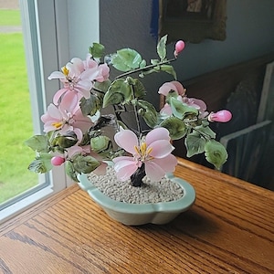 May include: A decorative bonsai tree with pink glass flowers and green leaves. The miniature tree is planted in a light green ceramic pot filled with small pebbles. The bonsai is placed on a wooden surface near a window.