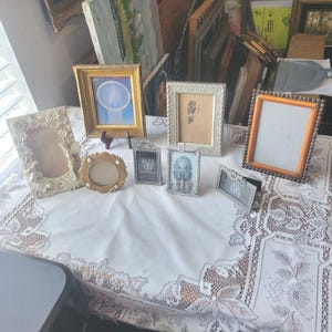 May include: A collection of decorative picture frames in various styles and sizes, arranged on a white lace tablecloth. Frames include gold, silver, and ornate designs, some with artwork or prints inside. The frames are of different shapes, including rectangular and oval.