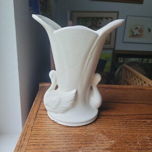 May include: A matte white ceramic vase featuring a flared, scalloped rim and a sculpted base adorned with two swan figures on opposite sides. The body of the vase has vertical ridges extending from the base toward the top opening.
